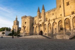 The Palais des Papes in Avignon viewed from the plaza in warm evening light with a few visitors for scale.